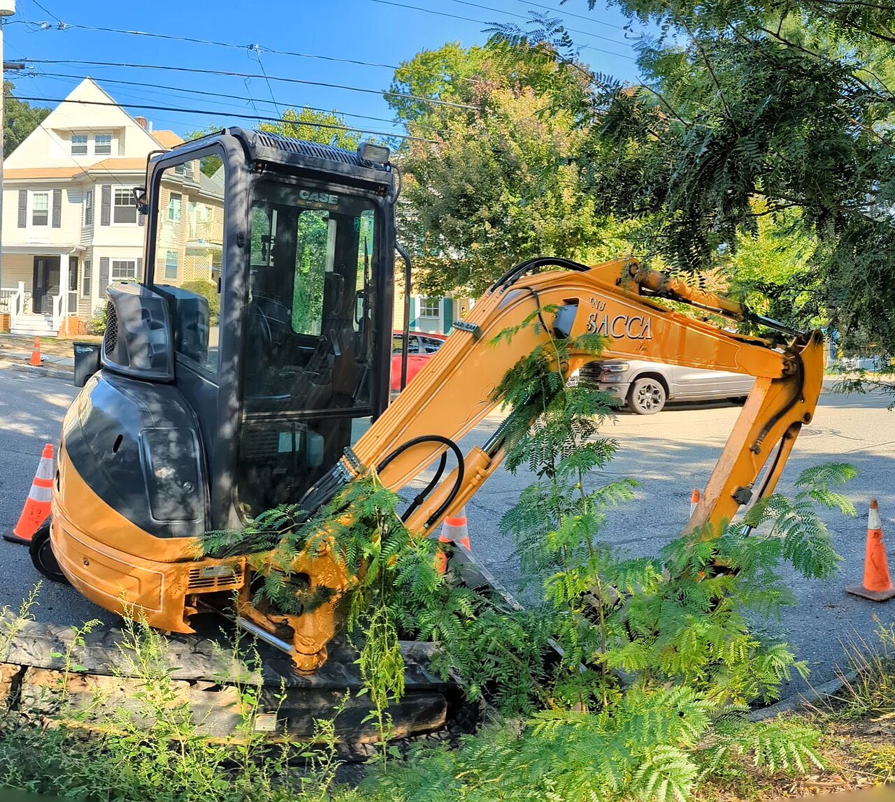 Compact Case excavator clearing brush on a tree-lined residential street